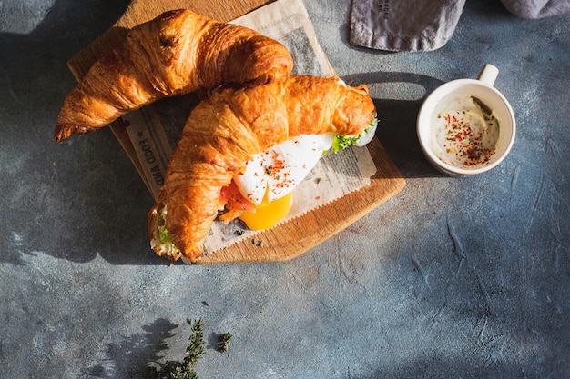 croissant sandwich with poached egg, salad leaves, salmon and cottage cheese seaved on cutting board with coffee. breakfast concept