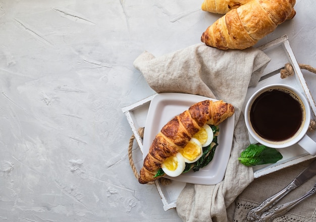 Croissant with egg and sauteed spinach and coffee in wooden tray on light gray concrete.