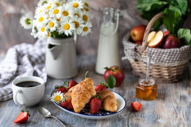 Croissants with fresh red strawberries with a Cup of coffee on a wooden table and bouquet of daisies in a jug.