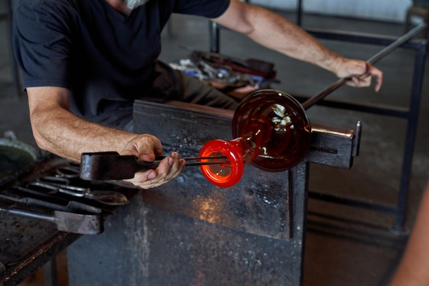 Crop anonymous male master using professional metal tool to shape incandescent glass vase while working in manufactory