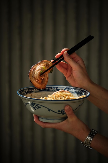 Crop anonymous person eating delicious crispy slice of chicken via chopsticks while holding ceramic bowl with ramen near metal wall