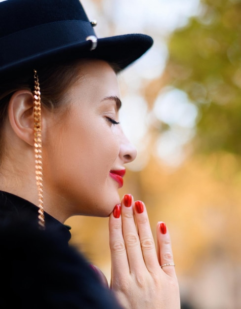 Crop of face of female model with natural makeup and bright red lips dressed in vogue hat with gold chain keeping hands together while closing eyes and dreaming of something on open air