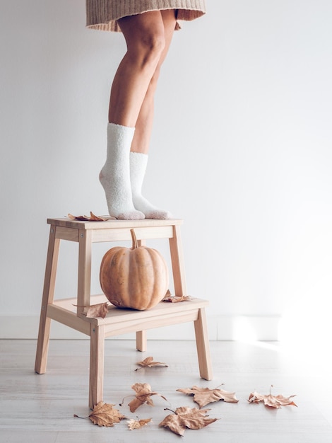 Crop legs of anonymous female in white socks standing on wooden stand decorated with ripe pumpkin and scattered dried autumn leaves against white wall in studio