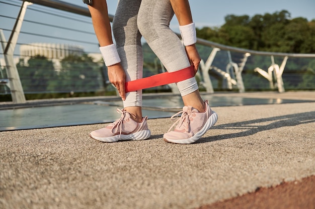 Crop of the legs of an unrecognizable female athlete exercising with resistance fitness band, doing body weight training outdoor on the city bridge. Weight loss, fitness and sport concept. Close-up.