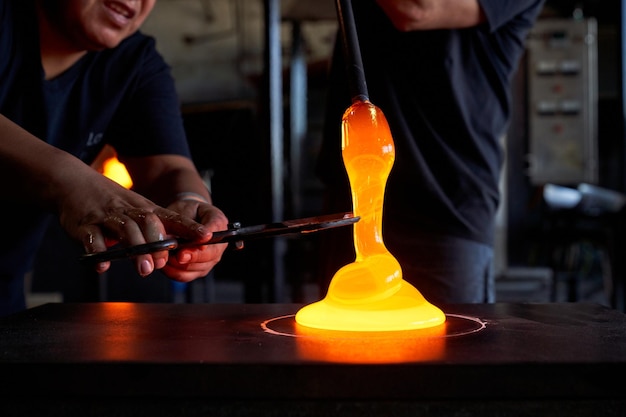 Crop male glassmiths in uniform in process of forming hookah vase using blowpipe and sheers while working at professional glassblowing factory together