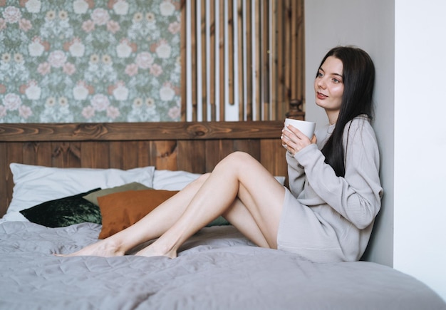 Crop photo of Beautiful smiling woman teenager girl with dark long hair in home clothes with morning cup of coffee on the bed