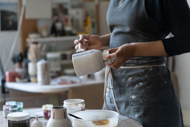 Crop photo of woman artisan making earthenware vase preparing for paint with brush