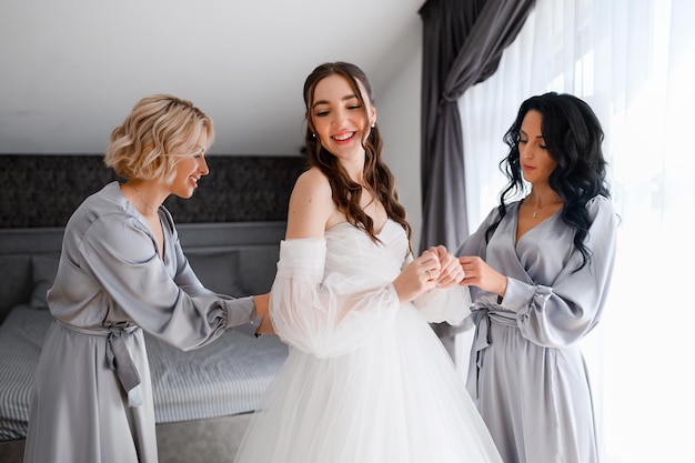 Crop portrait of pretty elegant woman bride in puffy white dress smiling while bridesmaids helping prepare for wedding ceremony standing spacious room in silk gray robe Morning of bride