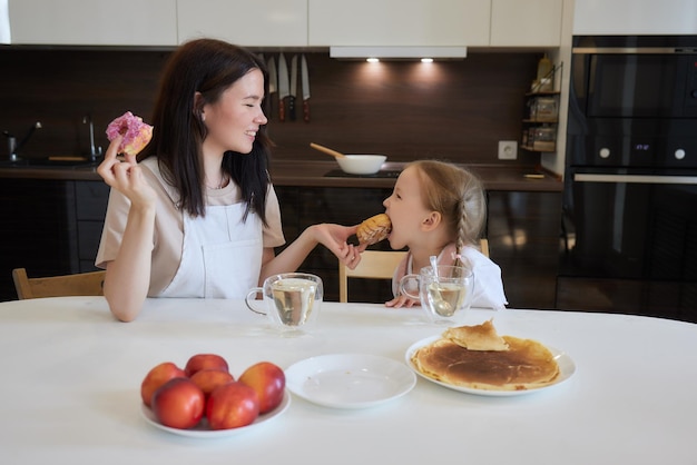 Crop shot of mother in red and her daughter having colorful donuts sitting at kitchen Dieting concept and junk food