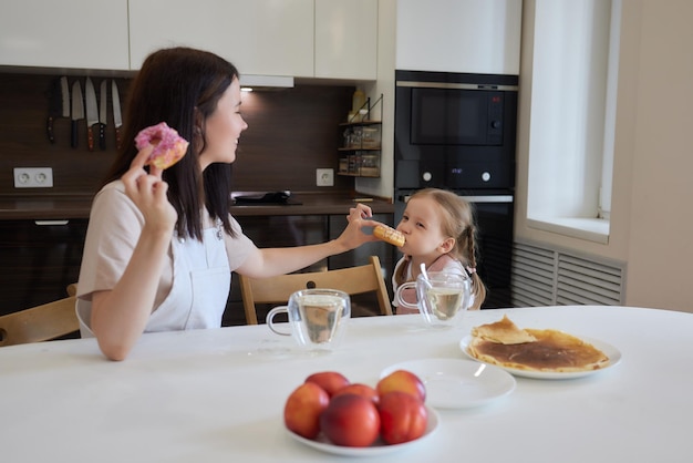 Crop shot of mother in red and her daughter having colorful donuts sitting at kitchen Dieting concept and junk food