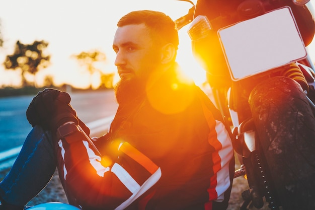 Crop side view of attractive bearded man in protective jacket sitting in evening at rear wheel of motorcycle on blurred backlit background of empty road