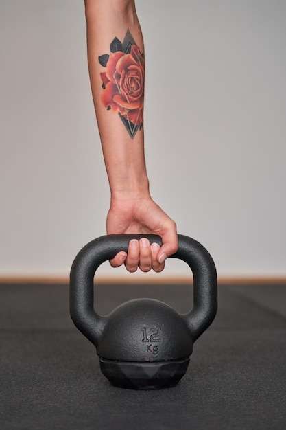 Crop unrecognizable female athlete with tattooed hand holding heavy kettlebell while preparing for workout in gym