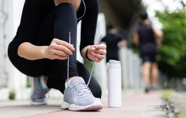 Crop unrecognizable woman runner wearing hygiene protective face mask sitting and tieing shoe rope during workout running in city park. Concept of healthcare in time of coronavirus outbreak.