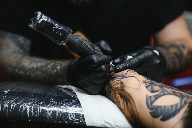 Cropped close up of a bearded tattoo artist working at his studio tattooing sleeve on the arm of his male client. Man getting tattooed by professional tattooist