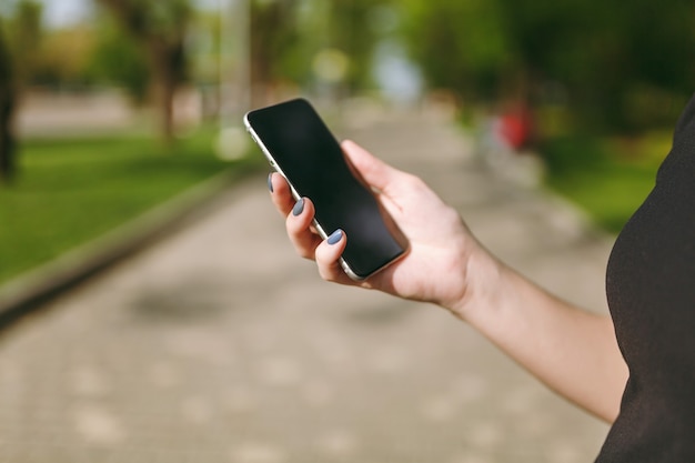 Cropped close up of woman hands holding and using mobile phone, smartphone with blank empty screen in city park outdoors