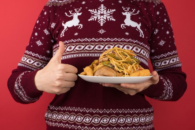 Cropped closeup photo of woman in red and white christmas sweater holding large plate of food and making thumbup on isolated red background