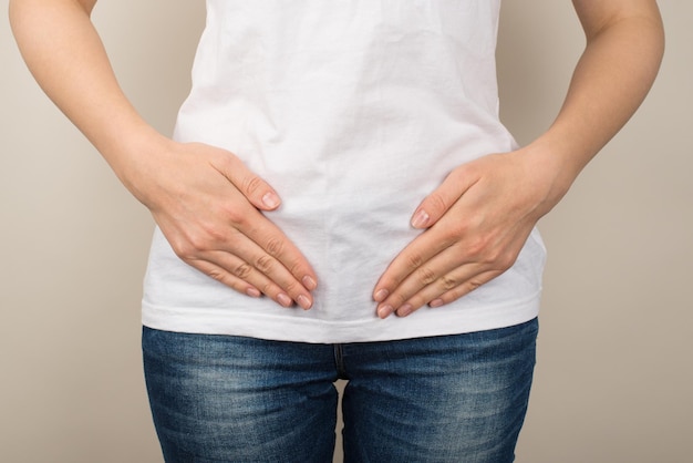 Cropped closeup photo of young woman in white tshirt and jeans holding her hands on lower abdomen on isolated grey background