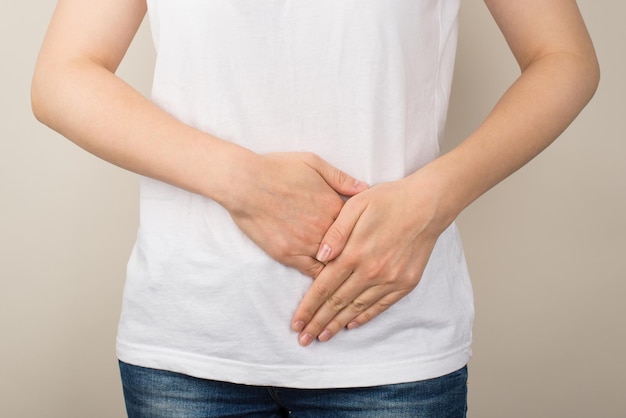 Cropped closeup photo of young woman in white tshirt and jeans holding left lower abdomen with her hands on isolated grey background