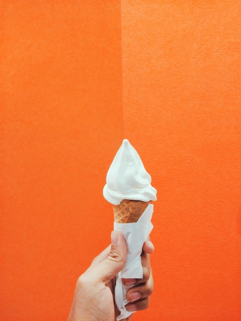 Cropped hand holding ice cream cone against orange wall