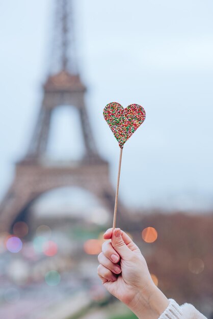 Photo cropped hand of woman holding heart shape candy against eiffel tower