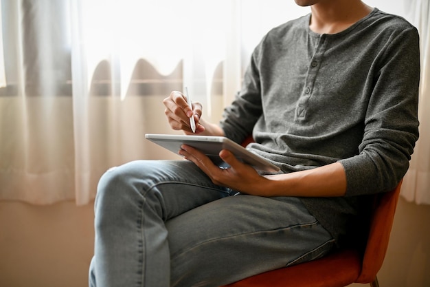 Cropped image of an Asian man using his digital tablet while relaxing on a chair in living room