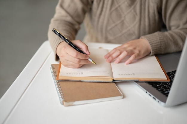 A cropped image of an Asian woman taking notes in her notebook while working on her laptop computer