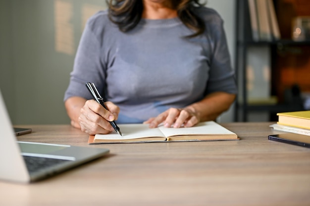 Cropped image of an Asianaged businesswoman taking notes on her planner book at her table