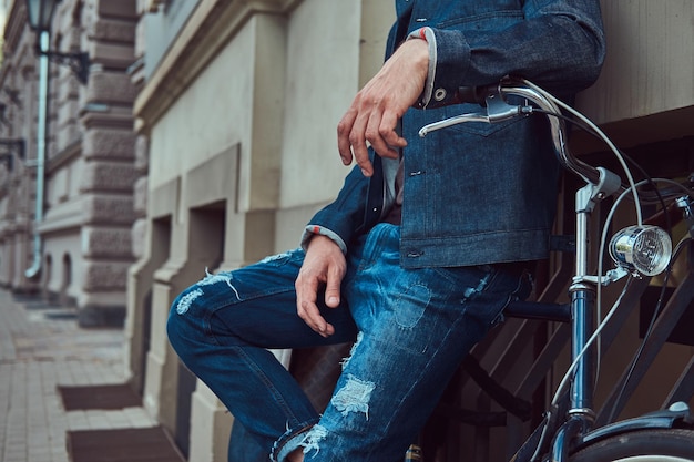 Cropped image of a fashionable man in stylish clothes leaning against a wall with city bicycle on a street.