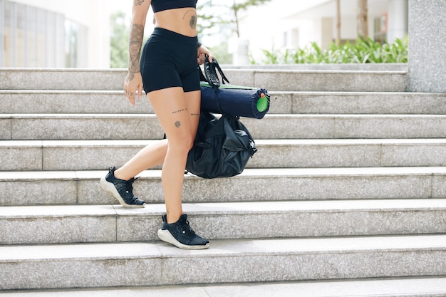 Cropped image of fit young woman in shorts and sports bra walking down the stairs with gym bag in hands