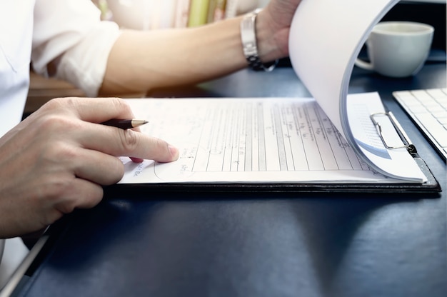 Cropped image of man holding pen and open clipboard with paper work while sitting at office desk.