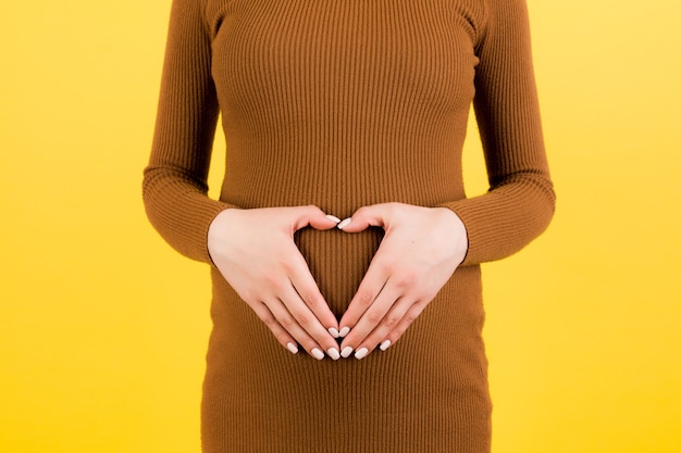 Cropped image of pregnant woman in brown dress making heart shape with the hands on her abdomen at yellow background. Baby bump concept. Copy space.