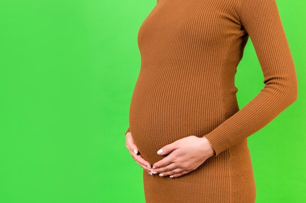 Cropped image of pregnant woman hugging her abdomen. Mother is wearing brown dress at green background. Future parent is expecting her child. Copy space.