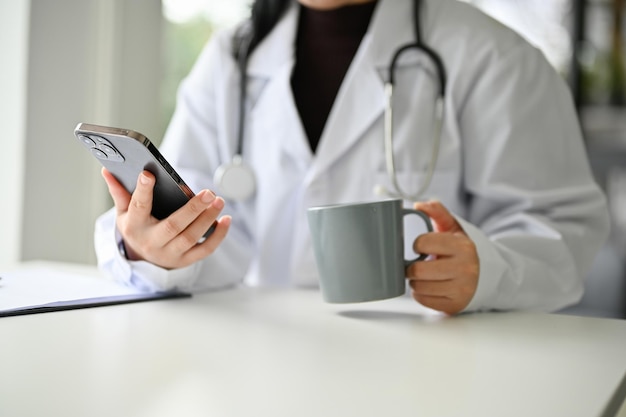 Cropped image of a relaxed Asian female doctor sipping coffee while using smartphone at her desk