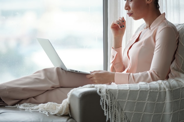 Cropped image of serious young woman sitting in armchair and reading document on laptop screen