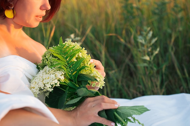 Cropped image of woman in a field with a bouquet of hydrangea in a white men's shirt at sunset in summer, beauty in nature concept