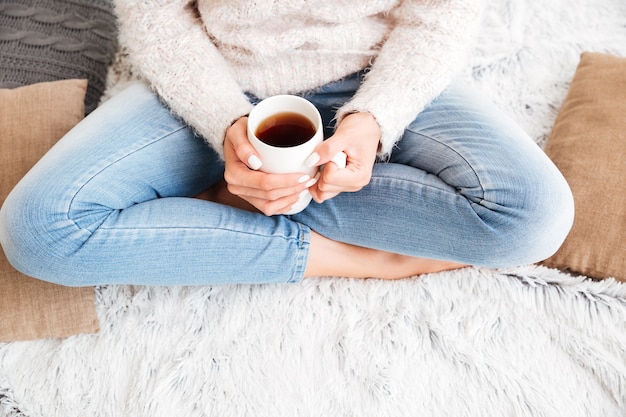 Cropped image of a woman in sweater and jeans holding tea cup while sitting on the carpet indoors