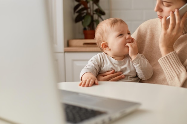 Cropped image woman wearing beige sweater sitting in kitchen and working online on laptop and talking via smartphone looking after baby girl calming down crying daughter