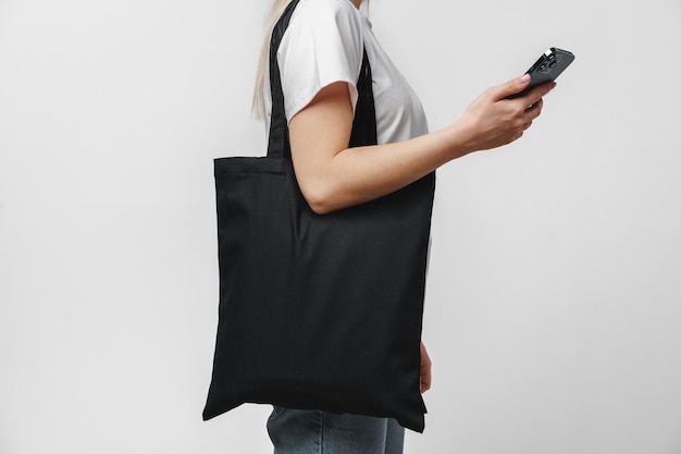 Cropped image of woman with black cotton shopping bag using smartphone against white background