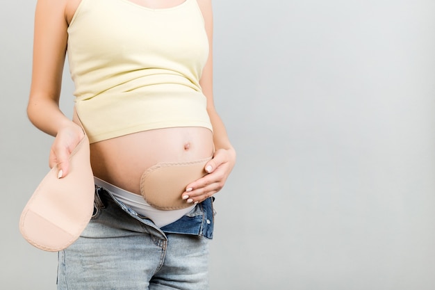 Cropped image of young pregnant woman dressing pregnancy bandage on her belly on gray surface. Copy space. Orthopedic abdominal support belt concept.