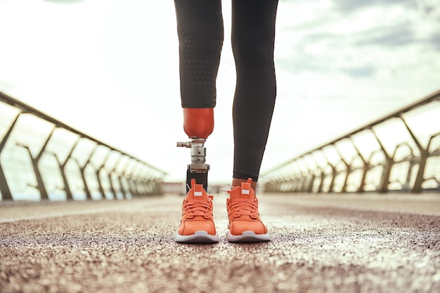 Cropped photo of disabled woman with prosthetic leg in sportswear standing on the bridge