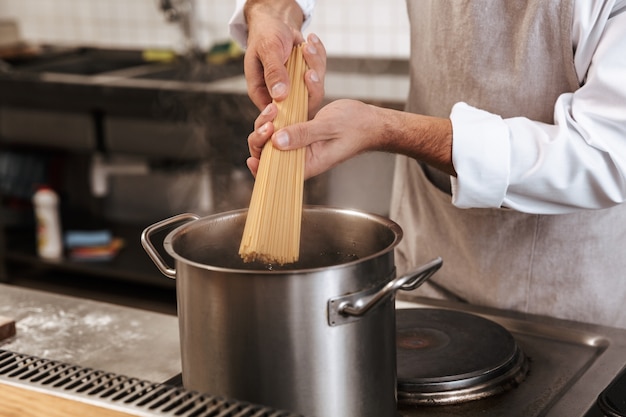 Cropped photo of european man chief in white uniform cooking pasta in pot, at kitchen in restaurant