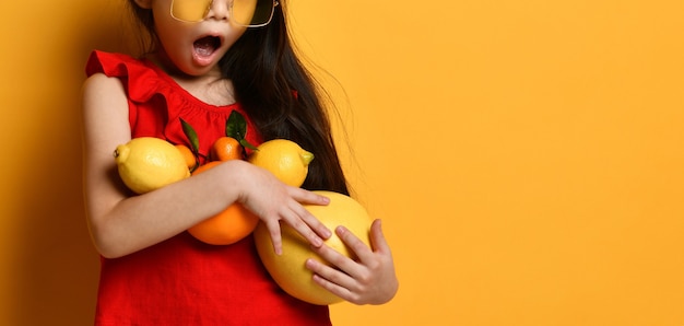 Cropped photo of an excited Little Asian girl in a red blouse holding pomelo, oranges and lemons in her hands, posing on an orange background. Childhood, fruits. Close copy space