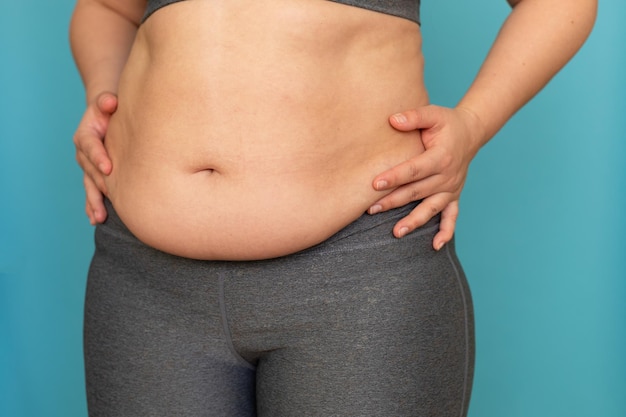 Cropped photo of fat woman in grey sports bra leggings showing excess belly putting hands on waist on blue background