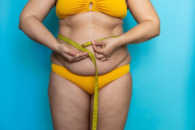 Cropped photo of plump woman standing in yellow swimming trunks showing excess naked belly measuring waist with tape