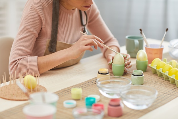 Cropped portrait of unrecognizable young woman painting eggs in pastel colors for Easter while sitting at table in kitchen , copy space