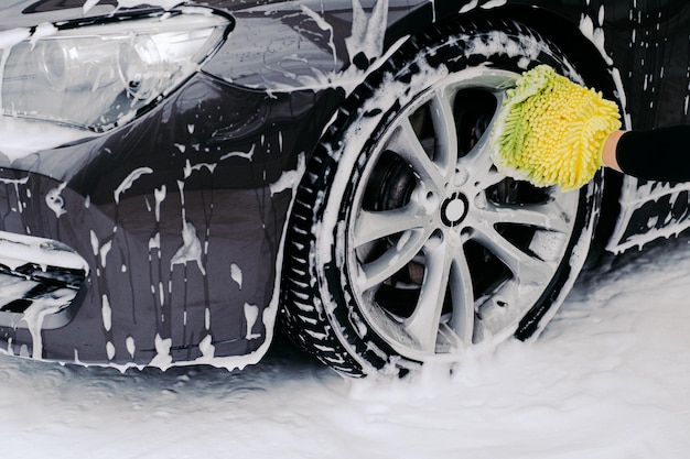 Cropped shot of black automobile washed by service worker at carwash Vehicle covered with foam bubbles Focus on wheel