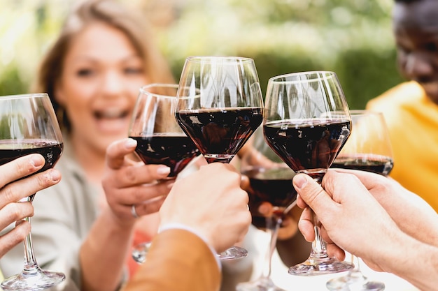 Cropped shot of friends toasting wineglasses of cabernet Selective focus on the glasses with the red wine blurred multiracial friends smiling on background alcohol lifestyle concept