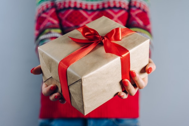 Cropped shot of girl in knitted sweater with traditional christmas pattern holding gift box wrapped in craft paper and decorated with red satin ribbon