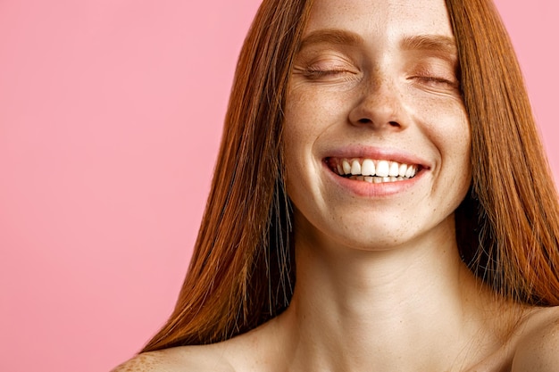 Cropped shot of happy caucasian woman with red shiny hair, clean freckled skin, bare shoulders, laughing with closed eyes, posing over pink background. Spa, skin care, beauty concept.