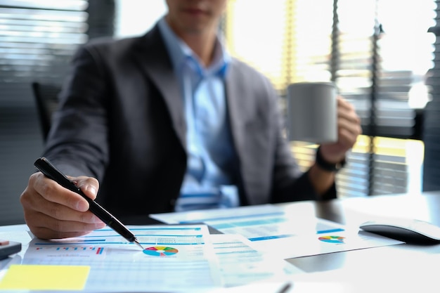 Cropped shot of male economist drinking coffee and working with sales statistics document at office desk